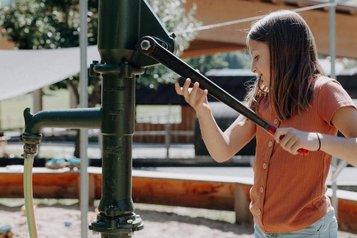 Ein Kind spielt am Wasserspielplatz mit einer Pumpe im Außenbereich des Familienhotels Oberkarteis im Salzburger Land.