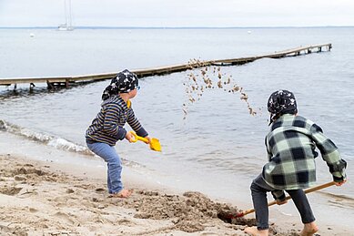 Zwei Kinder spielen mit dem Sand am Strand des Familienhotels Strandkind und werfen mit dem Sand um sich.