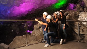Vier Personen mit Helmen stehen an einem Geländer in einer Höhle im Erlebnis Bergkwerk Merkers; ein Mann kniet und deutet auf leuchtende Kristallformationen in Lila, Blau und Grün.