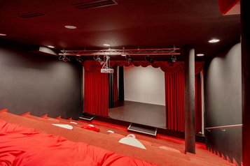 Blick auf eine kleine Bühne mit rotem Vorhang, davor gestufte Sitzflächen mit roten Sitzkissen in einem dunklen Raum im Familienhotel Amiamo.