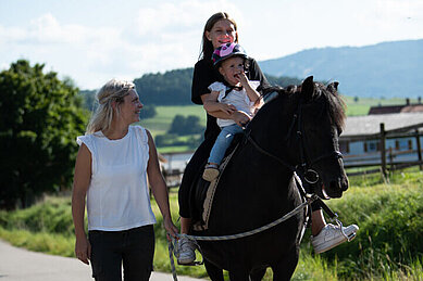 Zwei Kinder sitzen auf einem Pferd. Das Pferd wird von der Mutter geführt.