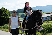 Zwei Kinder sitzen auf einem Pferd. Das Pferd wird von der Mutter geführt.