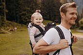 Papa trägt seine kleine Tochter in einer Trage auf den Rücken draußen in der umliegenden Natur des Familienhotels Habachklause spazieren.