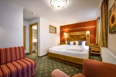 Gemütliches Doppelzimmer im Hotel Zauchenseehof mit warmen Rot- und Holztönen, großem Holzbett, klassischem Teppichboden, einladender Sitzgelegenheit und direktem Blick ins angrenzende Badezimmer.