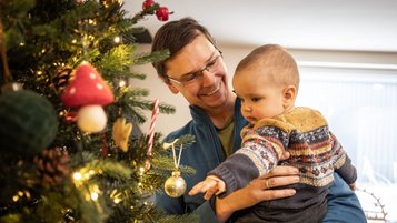 Vater hält sein Baby im Arm, während das Kind neugierig nach dem weihnachtlich geschmückten Christbaum im Familienhotel greift.