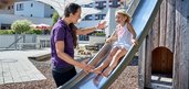 Kind rutscht eine Rutsche auf dem Outdoor Spielplatz des Familienhotels Amiamo hinunter. Eine freundliche Kinderbetreuerin hilft ihr dabei.
