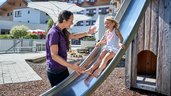 Kind rutscht eine Rutsche auf dem Outdoor Spielplatz des Familienhotels Amiamo hinunter. Eine freundliche Kinderbetreuerin hilft ihr dabei.