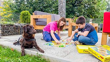 Kinder spielen im Garten des Familienhotels Engel ein Brettspiel. Ihr Hund liegt daneben.