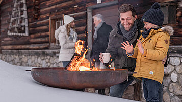 Winter im Salzburger Land: Familie steht vor einer Hütte, dick eingepackt in Winterklamotten. Papa und Kind wärmen sich an einer Feuerschale die Hände auf.