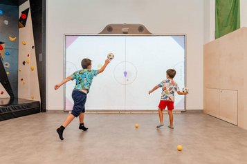 Zwei Jungen werfen Bälle auf eine große digitale Leinwand, auf der ein Spiel projiziert wird; links eine Boulderwand im Familienhotel Amiamo.