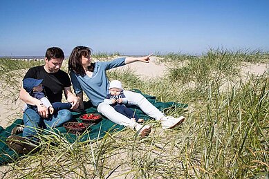 Eine Familie genießt ein Picknick auf einer grünen Decke in den Dünen mit Blick auf die Nordsee, wobei die Mutter auf etwas in der Ferne deutet, während der Vater und zwei kleine Kinder sich entspannen und Erdbeeren essen, was die familienfreundliche Umgebung und die Nähe des Familienhotels Deichkrone zum Strand hervorhebt.