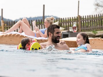 Vater mit seinen zwei Kindern im Aussen-Pool eines Hotels. Mutter liegt gemütlich auf einer Liege und liest ein Buch 