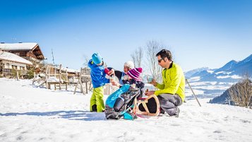 Eltern sitzen mit ihren beiden Kindern auf einem Schlitten im Schnee.