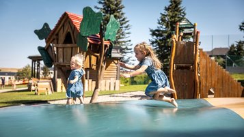Zwei kleine Kinder spielen auf einem Bodentrampolin vor einem Holzspielplatz im Freien.