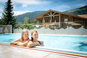 Zwei Mädchen lehnen am Beckenrand eines Pools, im Hintergrund ein Holzhaus und grüne Berge, im Familienhotel Spa- & Familien-Resort Krone im Allgäu.