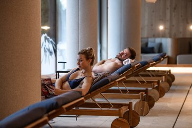 Entspannungsbereich im Zauchenseehof: Eine Frau liest auf einer gepolsterten Holzliege, während ein Mann auf der Liege dahinter ruht; große Säulen und bodentiefe Fenster schaffen eine ruhige Wellnessatmosphäre.