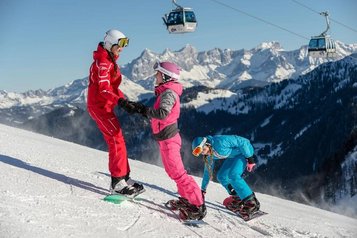 Snowboardkurs im Zauchenseehof mit Skilehrerin und zwei Kindern auf einer sonnigen Skipiste, im Hintergrund eine verschneite Berglandschaft mit Gondelbahn