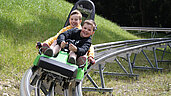 Zwei Jungs auf der Sommerrodelbahn "BocksBergBob" im Harz. Beide fahren mit strahlenden Gesichtern die Bahn hinunter.