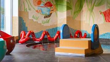 Bunter Indoor-Wasserbereich mit kleinem Becken, Mini-Rutsche, roten Wasserschlangen und verspieltem Wandbild für Kleinkinder im Familienhotel Amiamo.