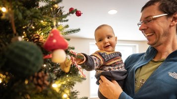Vater hält sein Baby im Familienhotel Elldus Resort auf dem Arm, während das Kind neugierig den geschmückten Weihnachtsbaum betrachtet.