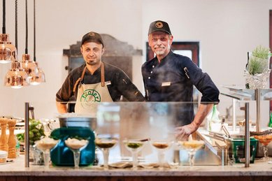 Zwei Köche des Familienhotels Tirolerhof stehen hinter der offenen Kochstation und lächeln in die Kamera.