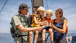 Familie mit zwei Kindern am Gipfel eines Berges in Tirol mit einem Gipfelbuch in der Hand.