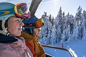 Zwei Teens sitzen im Winter im Sessellift und fahren die Skipiste hinauf.