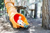 Kind rutscht aus einer gelben Röhrenrutsche im Waldspielplatz des Familienhotels Huber in Südtirol