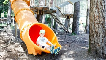 Kind rutscht aus einer gelben Röhrenrutsche im Waldspielplatz des Familienhotels Huber in Südtirol