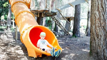 Kind rutscht aus einer gelben Röhrenrutsche im Waldspielplatz des Familienhotels Huber in Südtirol