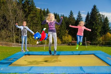 Drei Kinder springen lachend auf einem großen Outdoor-Trampolin im Familienhotel Ramsi in Kärnten inmitten grüner Natur
