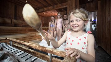 Mädchen gießt in der Sauna eines Familienhotels Wasser auf die Steine, während die Familie im Hintergrund gemeinsam lacht.