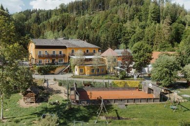 Das Kinderhotel Bruckwirt in Oberösterreich ist in einer Drohnenaufnahme mit Fluss im Vordergrund zu sehen.