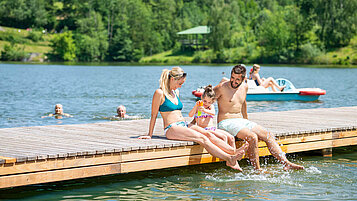 Familie sitzt auf dem Badesteg am Eginger see und lässt die Füße ins Wasser hängen.