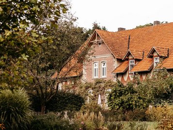 Rotes Backsteinhaus mit orangefarbenem Ziegeldach, umgeben von dichtem Garten mit blühenden Stauden und Büschen.