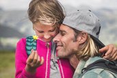 Vater und Tochter genießen im Familienhotel Egger eine innige Umarmung bei einer Wanderung in der Natur, während das Mädchen eine gelbe Blume betrachtet.