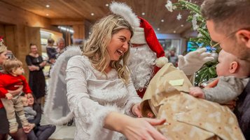 Eine Frau als Engel verkleidet und der Weihnachtsmann überreichen einem Baby in den Armen seines Vaters ein Geschenk während einer fröhlichen Adventsfeier im Familienhotel Das Bayrischzell