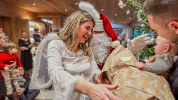 Eine Frau als Engel verkleidet und der Weihnachtsmann überreichen einem Baby in den Armen seines Vaters ein Geschenk während einer fröhlichen Adventsfeier im Familienhotel Das Bayrischzell