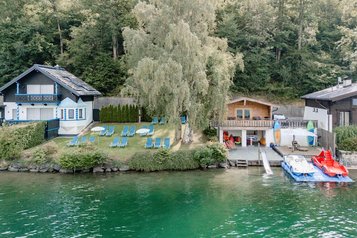 Liegewiese mit Sonnenliegen, Badeplattform mit Rutsche und zwei Tretbooten an einem klaren See, umgeben von Bäumen.