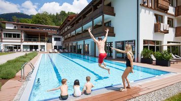 Familie am Außenpool eines Alpenhotels, während ein Mann ins Wasser springt.