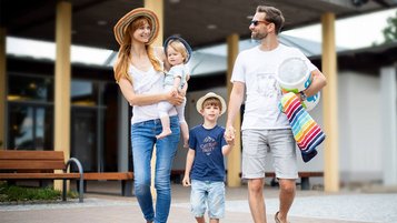 Familie mit zwei Kindern in Sommerkleidung auf dem Weg zum Schwimmbad, mit Handtuch und Schwimmring.