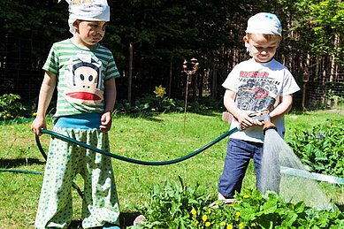 Zwei als Piraten verkleidete Kinder stehen im Gemüsegarten des Familienhotels Borchard's Rookhus an der Mecklenburgischen Seenplatte und gießen mit einem Gartenschlauch die Pflanzen.