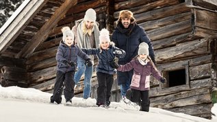 Familie tobt fröhlich im Schnee vor einer urigen Holzhütte und genießt den Winterurlaub im Familienhotel.