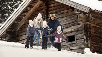 Familie tobt fröhlich im Schnee vor einer urigen Holzhütte und genießt den Winterurlaub im Familienhotel.