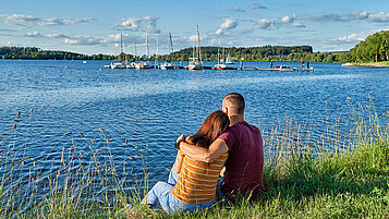 Fichtelgebirge im Sommer. Verbringt schöne Stunden an den Seen. Ein paar sitzt am Ufer. Im Hintergrund sind Segelboote an einem Steg angelegt. 