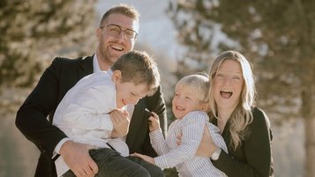 Gastgeberfamilie mit zwei Kindern lacht und posiert fröhlich vor winterlicher Bergkulisse beim Familienhotel Alpenhof Dolomit Family.