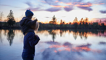 Mutter steht mit ihrem Kleinkind auf den Schultern am Seeufer des Chiemsees und betrachtet die abendliche Stimmung.