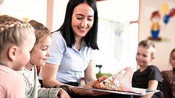 Drei Kinder in der Kinderbetreuung mit einer Kinderbetreuerin. Die Betreuerin liest ein Buch vor.