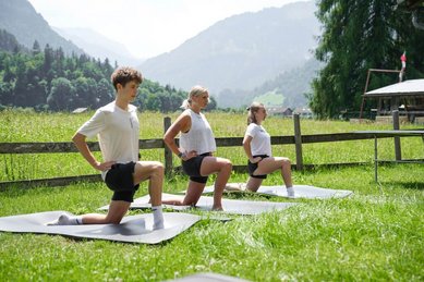 Drei Personen machen Yogaübungen auf Matten im Freien mit Blick auf die Berge beim Familienhotel Sonne Bezau.