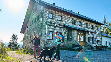 Der Berggasthof Dreisesse im Bayerischen Wald steht im Hintergrund von zwei Wanderern mit Hund.